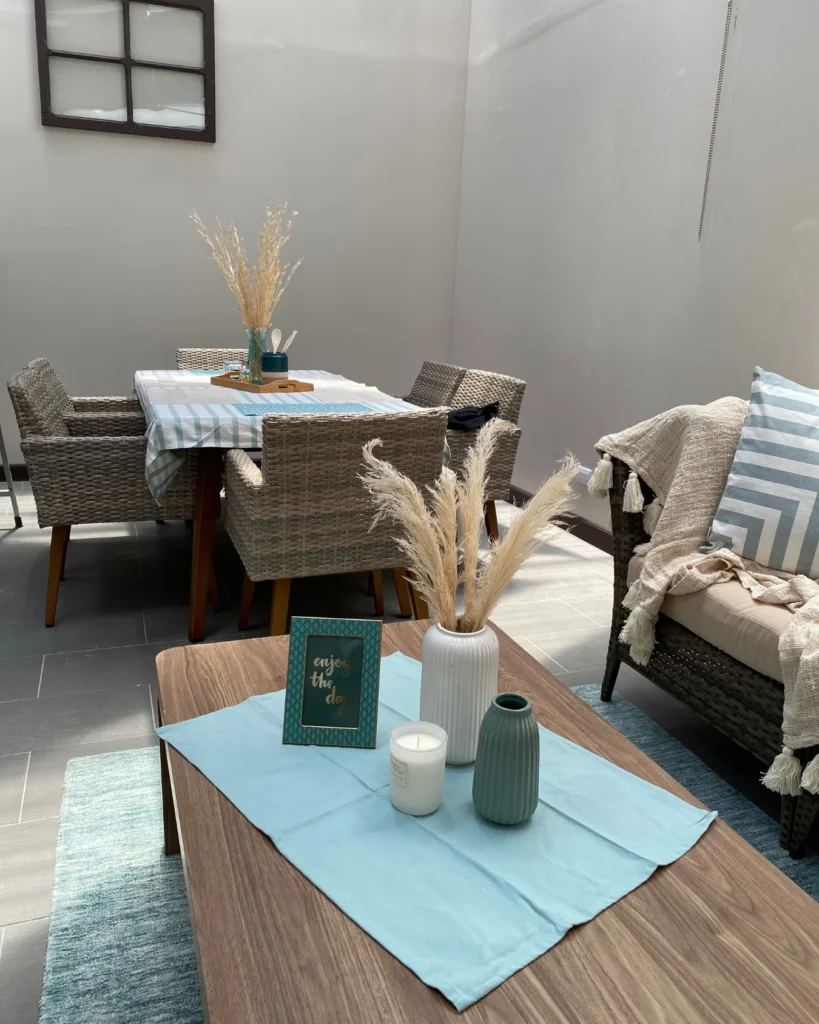 Área de comedor con sillas de fibra y centro decorativo; en primer plano mesa de sala con jarrones y vela, tonos azul y beige.