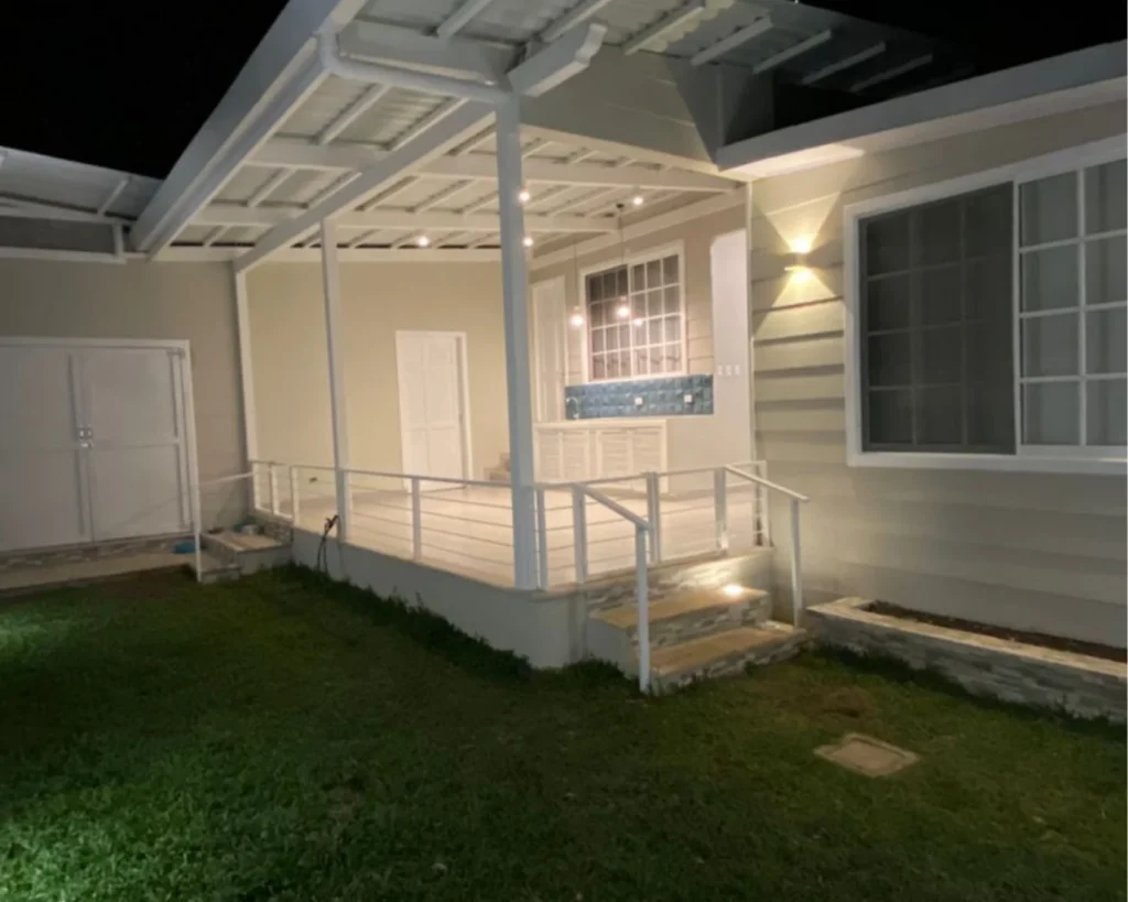Terraza techada de casa restaurada en Escazú, con barandales blancos, iluminación nocturna y detalles modernos en revestimiento y ventanería.
