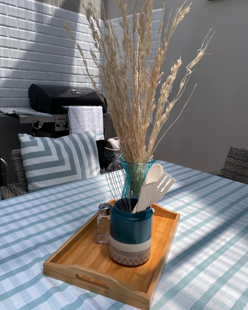 Primer plano de bandeja en mesa con jarrón, utensilios y salero; al fondo parrilla y pared de cerámica tipo subway en la terraza interna.