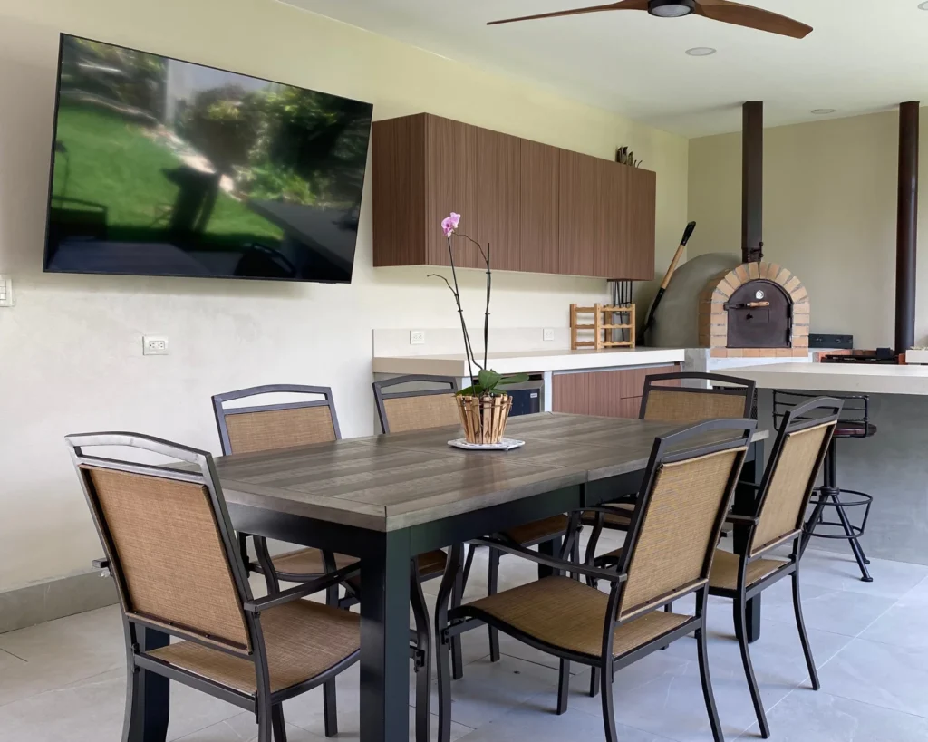Área de comedor con mesa y sillas, televisión mural y horno de pizza al fondo, dentro de la terraza techada remodelada