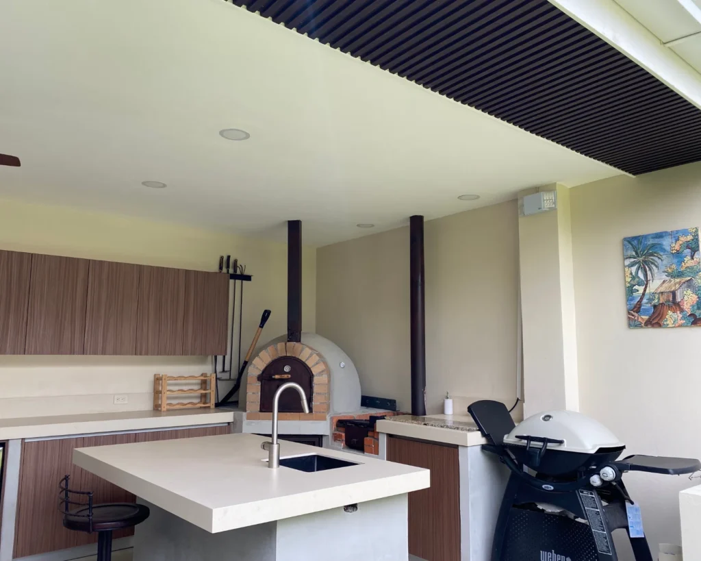 Vista completa de la cocina exterior terminada, con horno de leña, área de barbacoa, fregadero y mueble superior de madera