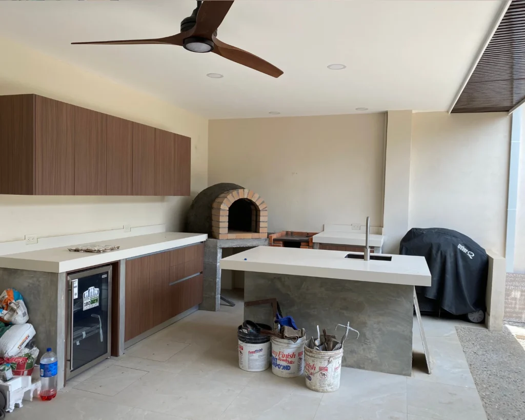 Cocina exterior con horno de leña, isla en concreto con fregadero y muebles empotrados de madera, en etapa final de instalación