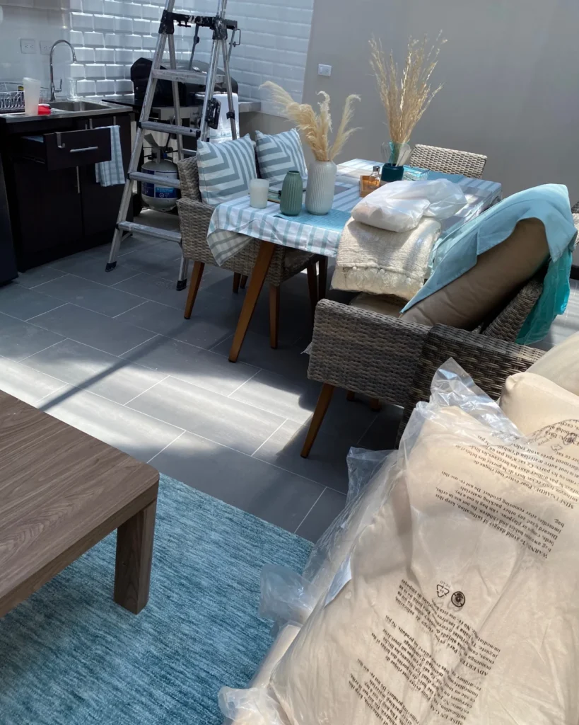 Vista del montaje: mesa con sillas tejidas, cojines y jarrones, escalera junto a fregadero y alfombra azul en la terraza del tercer piso.