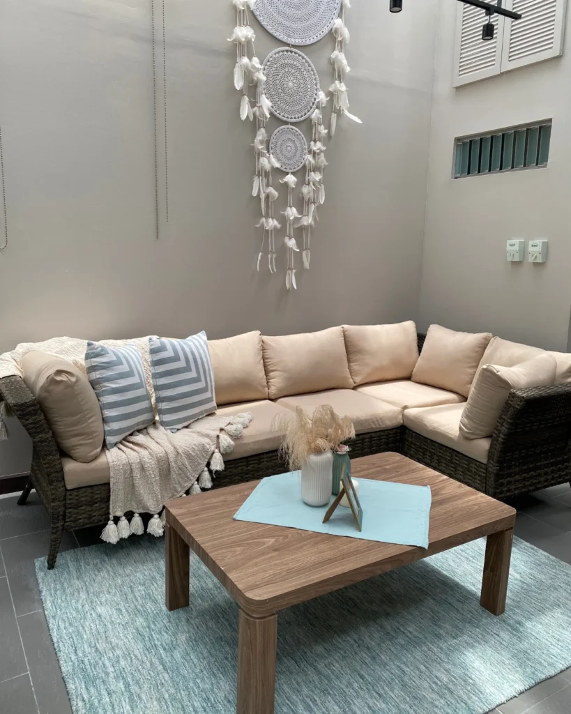 Sofá esquinero de ratán con cojines beige, mesa de centro sobre alfombra azul y tapiz decorativo tipo atrapasueños en pared, en terraza interna de clínica en Guadalupe.