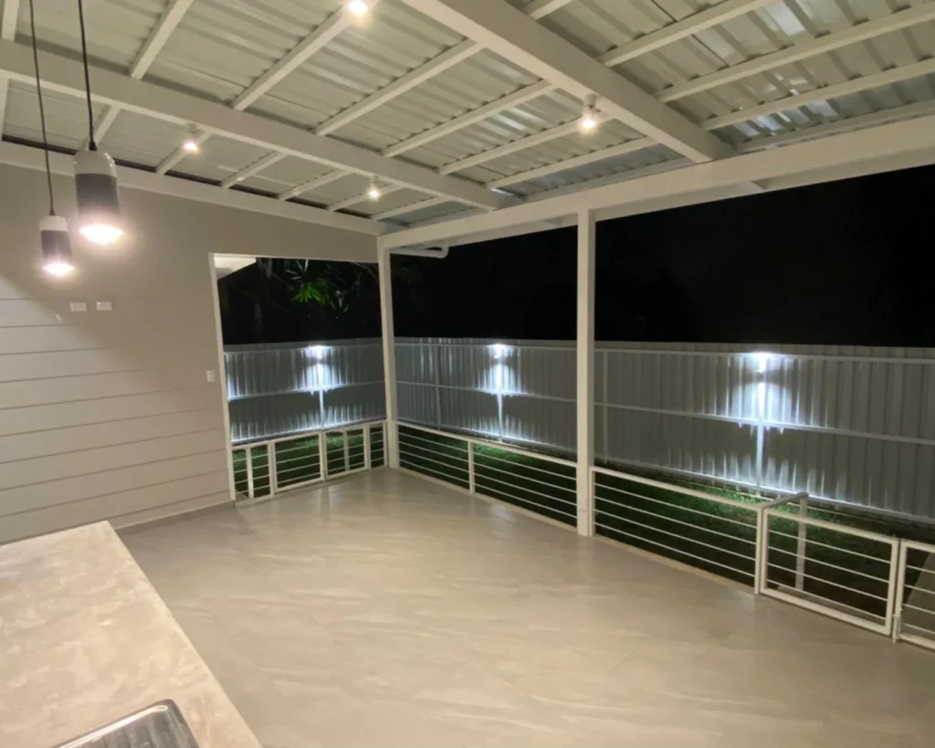 Terraza trasera techada con estructura metálica blanca, piso gris y luminarias empotradas; vista desde el área de servicio.