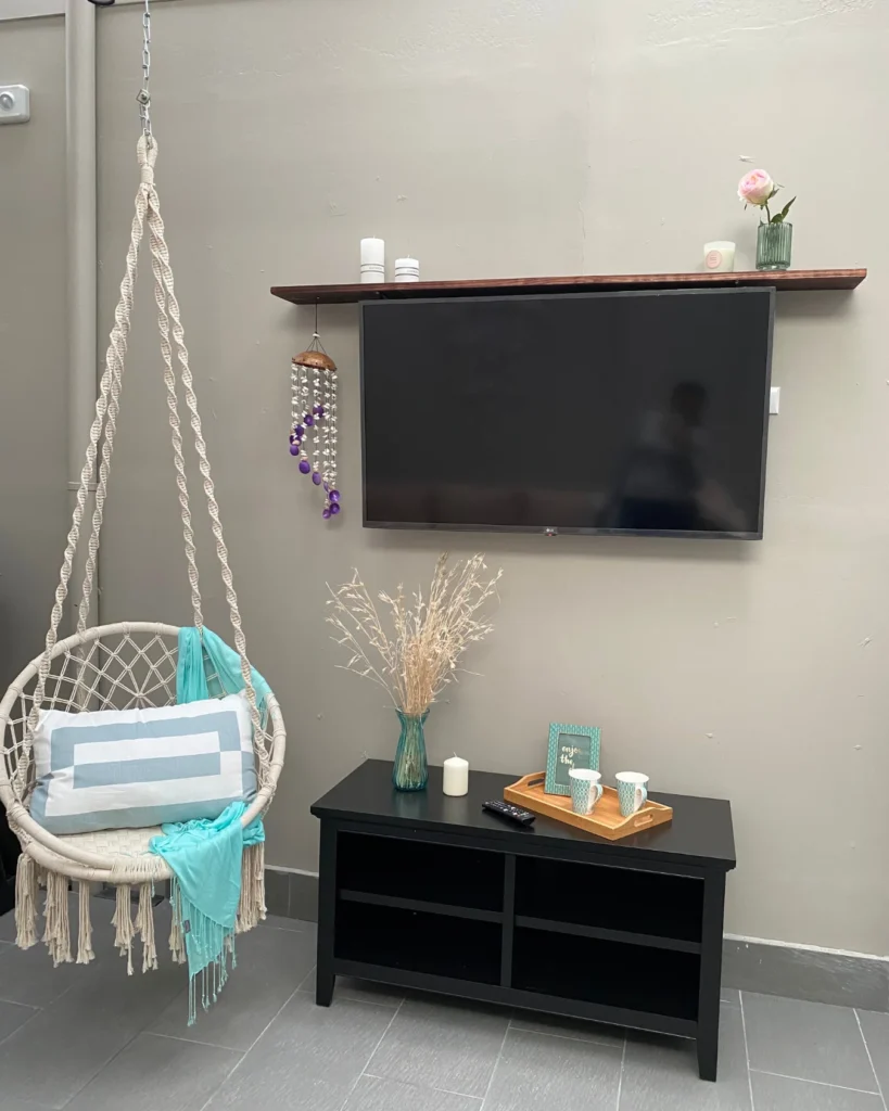 Zona de descanso con silla colgante de macramé, televisor en pared y mesa baja negra con bandeja y tazas.